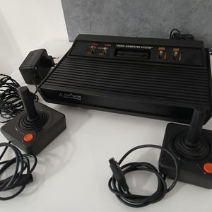 Könnte beinhalten: Schwarze Atari 2600 Videospielkonsole mit zwei Joysticks und Netzteil. Auf der Konsole steht "VIDEO COMPUTER SYSTEM". Die Joysticks haben orangefarbene Knöpfe.