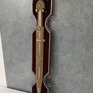 Peut inclure: Un poignard orné avec une lame et un fourreau dorés, monté sur une plaque de bois rouge foncé. La plaque est encadrée de métal argenté et porte le mot "GEORGIA" en haut. Le poignard présente des motifs décoratifs complexes.