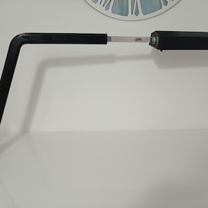 Może przedstawiać: Czarna lampa biurkowa z prostokątną głowicą i zakrzywionym ramieniem. Lampa stoi na białej powierzchni, w tle widoczny jest zegar. Design lampy jest minimalistyczny i nowoczesny.