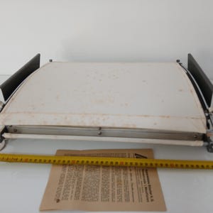 Könnte beinhalten: Antike Zeichenmaschine mit weißer Oberfläche und Metallrahmen. Die Maschine hat schwarze Seitenteile und einen schwarzen Knopf. Ein gelbes Maßband und ein Dokument befinden sich im Vordergrund. Die Maschine ist ca. 50 cm lang.