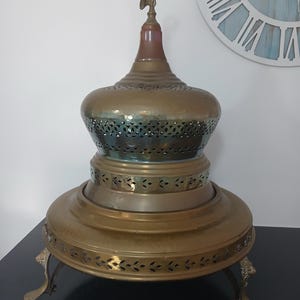 Può includere: Un scaldavivande antico in ottone con un design traforato decorativo. Lo scaldavivande ha una base rotonda sostenuta da gambe ornate, un corpo bulboso e un coperchio sormontato da una piccola statuina.