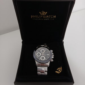 Op de afbeelding: Een zilverkleurig Philip Watch chronograaf horloge met een zwarte wijzerplaat en drie sub-wijzerplaten, gepresenteerd in een zwarte doos. Het horloge heeft een roestvrijstalen band en wordt gepresenteerd in een open, donkerbruine doos met de merknaam en het logo.