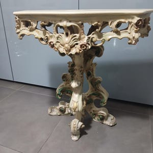Può includere: Una consolle ornata, color crema, con piano rettangolare e dettagli intagliati di fiori e volute. Il tavolo ha un supporto centrale con base decorativa ed è alto circa 76 cm.