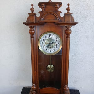Könnte beinhalten: Eine Vintage-Wanduhr aus Holz mit Pendel. Die Uhr hat ein rundes Zifferblatt mit schwarzen römischen Ziffern und goldenen Zeigern. Der Holzrahmen der Uhr ist dunkelbraun und hat dekorative Schnitzereien oben und unten.
