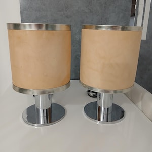 Puede incluir: Dos lámparas de mesa a juego. Cada lámpara tiene una pantalla cilíndrica beige con ribete plateado en la parte superior e inferior. La base es de metal brillante cromado con una base circular. Las lámparas están sobre una superficie blanca.