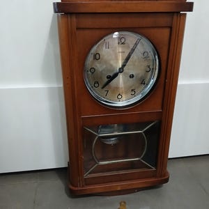May include: A vintage wooden wall clock with a round face and a pendulum window. The clock has a dark brown finish and a key is lying on the floor. The clock face has black hands and numerals.