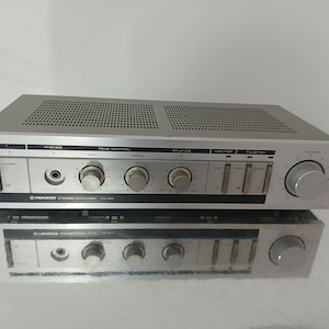Könnte beinhalten: Zwei silberne Pioneer Stereo-Verstärker übereinander gestapelt. Der obere Verstärker hat eine perforierte Oberseite, Knöpfe und Schalter. Der untere Verstärker spiegelt das Design wider. Beide Geräte zeigen das Pioneer-Logo. Ca. 43 cm breit.