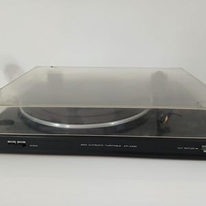 Może przedstawiać: Czarny automatyczny gramofon Akai AP-A100 33 RPM z przezroczystą plastikową osłoną przeciwpyłową. Gramofon posiada czarny talerz, ramię i panel sterowania. Logo Akai jest widoczne z przodu.