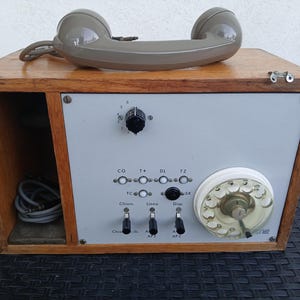 Può includere: Telefono vintage con cornetta grigia appoggiata su una scatola di legno. La scatola ha una facciata grigia con quadranti, pulsanti e un quadrante rotante. Il design del telefono ricorda la tecnologia della metà del secolo.
