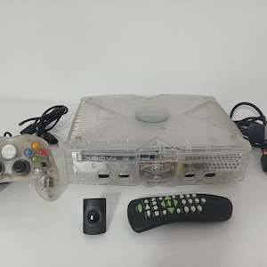 Könnte beinhalten: Eine transparente Xbox-Konsole mit passendem Controller, Fernbedienung und Zubehör. Das transparente Design der Konsole zeigt ihre internen Komponenten. Der Controller hat farbige Tasten. Eine schwarze Fernbedienung und ein kleiner schwarzer Empfänger sind ebenfalls enthalten.