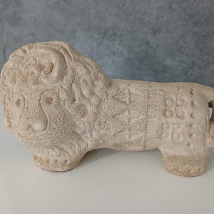 May include: A beige, handcrafted lion figurine. The sculpture has a textured surface and is decorated with carved patterns on its body. The lion is depicted in a standing position with its tail curled. The figurine is made of a light-colored material.