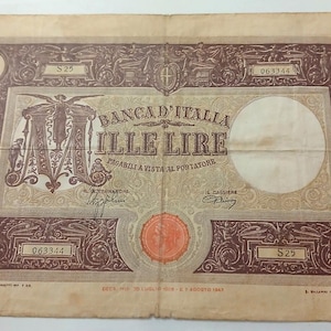 Può includere: Un'antica banconota italiana da 1000 lire. La banconota presenta volute ornate, il testo "BANCA D'ITALIA ILLE LIRE" e i numeri "1000" e "063344". La banconota ha un sigillo circolare e firme.