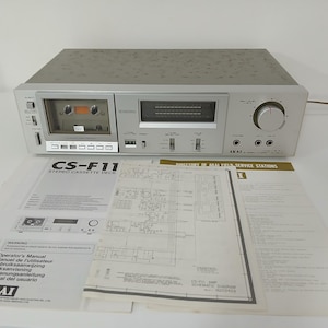 Może przedstawiać: Srebrny odtwarzacz kaset stereo AKAI CS-F11 z kasetą widoczną w odtwarzaczu. Odtwarzacz posiada szereg przycisków, pokręteł i wyświetlacz. Instrukcje i schematy znajdują się przed odtwarzaczem.