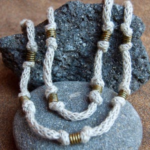 Puede incluir: Un collar hecho a mano con un cordón trenzado de color crema y detalles metálicos color latón. El collar se exhibe sobre una piedra gris, resaltando su estética natural y terrosa. El diseño incorpora nudos y anillos metálicos enrollados.