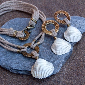 Può includere: Un set di gioielli composto da una collana e orecchini con dettagli in conchiglia naturale e ottone martellato. La collana è realizzata con una corda spessa naturale e ha una chiusura in ottone. Gli orecchini sono realizzati con cerchi in ottone con un ciondolo a conchiglia.