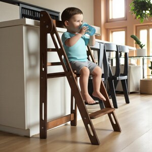 Può includere: Uno sgabello in legno marrone scuro con un design a scala. Lo sgabello ha più gradini e una piattaforma superiore. Un bambino è seduto sullo sgabello, che beve da una tazza blu. Lo sgabello è vicino a un piano di lavoro bianco.