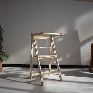 May include: A light-coloured wooden learning tower featuring a foldable design with a top horizontal bar, a middle support rail, a flat platform, and a bottom step. The structure is constructed from smooth, natural wood with visible grain patterns.