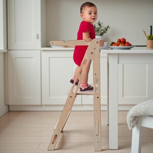 May include: A natural wood-colored toddler step stool with a safety bar. A child in a red outfit stands on the stool, looking towards the camera. The stool is positioned near a white table with a plate of tomatoes.
