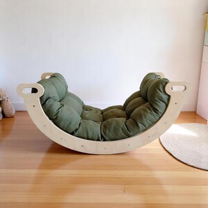 May include: A wooden rocking arch with a green, tufted cushion. The arch has handles on each side and is designed for children's play. The cushion is olive green and fills the curved space of the rocker.