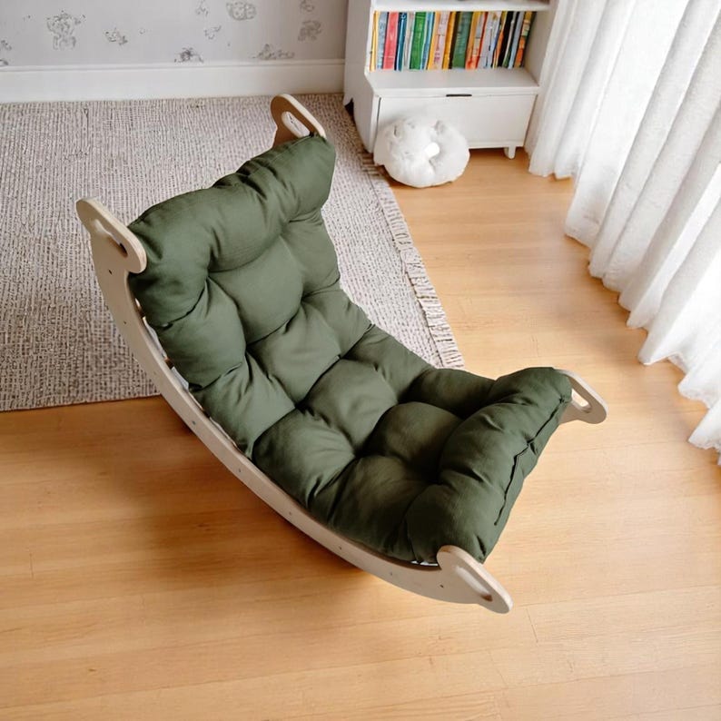 May include: A wooden rocking chair with a green cushion. The chair has a curved base and a padded seat and backrest. The cushion is a solid olive green colour. The chair is designed for comfort and relaxation.