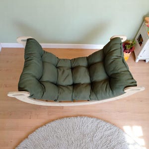 May include: A green, padded pet bed with a rocking base. The bed has a curved, wooden frame and a soft, tufted cushion. The bed is designed for small pets and is placed on a wooden floor.