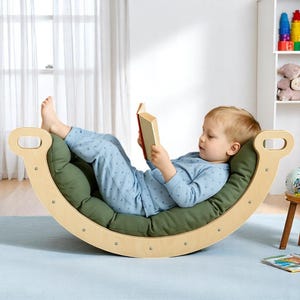 May include: A wooden rocking chair with a green cushion. A child in blue pyjamas is lying on the cushion, reading a book. The chair has a curved design with handles on each end.