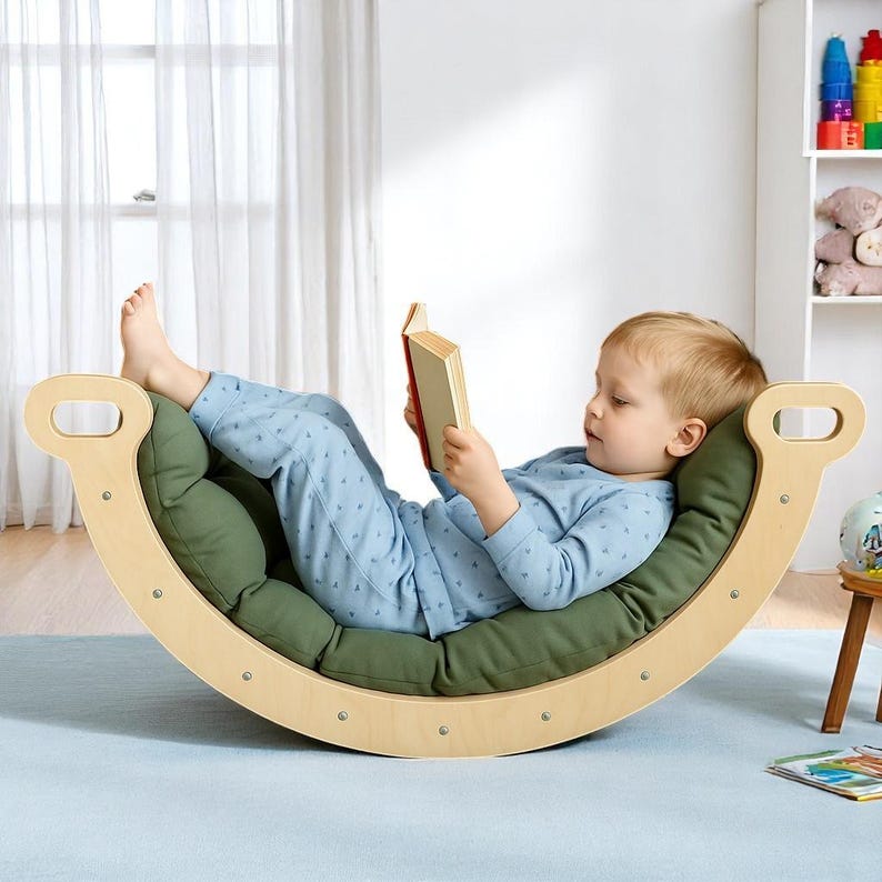 May include: A wooden rocking chair with a green cushion. A child in blue pyjamas is lying on the cushion, reading a book. The chair has a curved design with handles on each end.