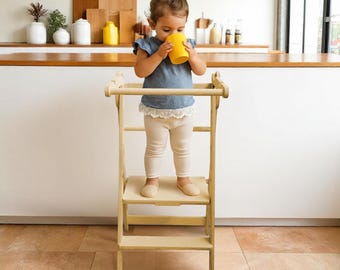 以下が含まれることがあります： 明るい木製の学習タワーに子供が立っています。子供は青いシャツとクリーム色のレギンスを着て、黄色のカップから飲んでいます。キッチンには木製のカウンターがあります。