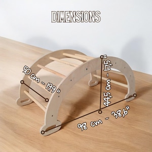 May include: A wooden climbing arch with dimensions displayed. The arch measures 98 cm long, 50 cm wide, and 44.5 cm tall. The text "DIMENSIONS" is at the top. The arch features several wooden rungs for climbing.