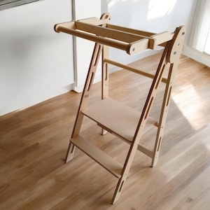 May include: A natural wood-toned learning tower with a step and a top rail. The tower has a ladder-like design with a platform for standing. The wood grain is visible, and the tower is designed for children's activities.