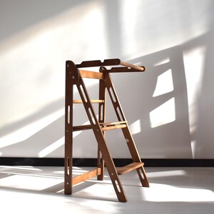 Könnte beinhalten: Ein Holztritt mit einem leiterartigen Design. Der Hocker ist aus hellbraunem Holz gefertigt und hat drei Stufen. Die oberste Stufe hat einen gebogenen Griff. Der Hocker steht vor einer weißen Wand.