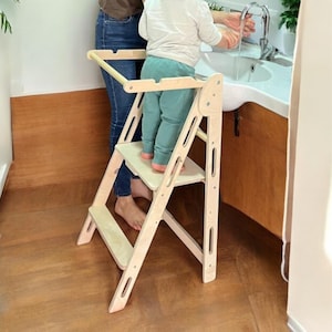 Könnte beinhalten: Ein natürlicher, zweistufiger Lernturm aus Holz mit Sicherheitsbügel. Der Turm wurde entwickelt, um Kindern zu helfen, Arbeitsplatten und Waschbecken zu erreichen. Ein Kind steht auf der oberen Stufe und wäscht sich die Hände an einem weißen Waschbecken.