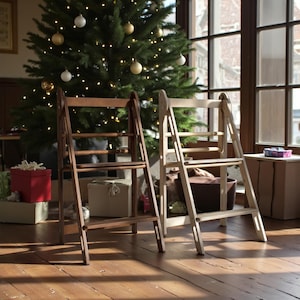 Può includere: Due scale a pioli pieghevoli in legno, una marrone scuro e una beige chiaro, sono su un pavimento in legno. Un albero di Natale decorato con ornamenti e luci è sullo sfondo, insieme a regali confezionati.