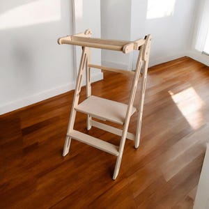 Può includere: Uno sgabello in legno chiaro con una barra superiore e una piccola piattaforma. Lo sgabello ha un design a scala con più pioli e una base robusta. Il legno ha una finitura naturale e lo sgabello è posto su un pavimento in legno.