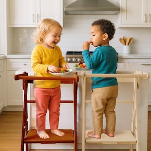 May include: Two children stand on wooden step stools in a kitchen, each holding a plate of fruit. One stool is dark brown, the other is light wood. The children wear colorful tops and pants. The kitchen has white cabinets and a stainless steel range hood.