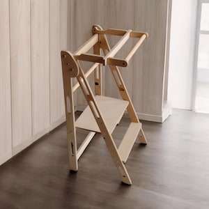 Può includere: Uno sgabello in legno chiaro con un ripiano e una barra superiore per appendere oggetti. Lo sgabello ha un design a scala con gambe angolate e un ripiano rettangolare. Il legno ha una finitura naturale.