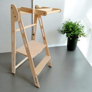 Può includere: Una torre di apprendimento in legno naturale con un gradino, un piccolo ripiano e una barra superiore. La torre ha un design a scala con lati aperti ed è progettata per i bambini. Una piccola pianta verde in un vaso nero è sullo sfondo.