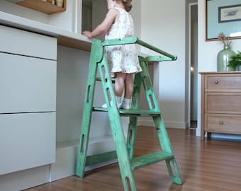 以下が含まれることがあります： 安全バー付きの緑色の木製ステップスツール。ステップスツールは、2つのステップとプラットフォームを備えたはしごのデザインです。スツールは使い古した緑色に塗装されています。子供がプラットフォームに立っています。
