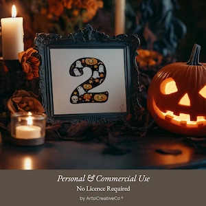 May include: A black framed picture with a white background and the number "2" in black with a Halloween theme. The number is surrounded by ghosts, pumpkins, and stars. The picture is sitting on a table with a lit jack-o'-lantern and candles.