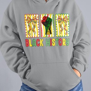May include: A gray hoodie with a front pocket and a graphic design. The design includes the text "BLACK HISTORY" in colorful letters, along with images of a raised fist and decorative bows.