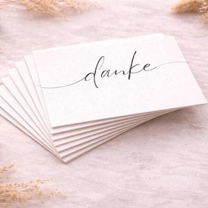 Könnte beinhalten: Ein Stapel weißer Karten mit dem Wort "danke" in eleganter schwarzer Schrift. Die Karten liegen auf einer hellrosa Oberfläche, mit getrockneten Pflanzen und einem kleinen Holzobjekt im Hintergrund. Die Karten sind wahrscheinlich für einen besonderen Anlass.