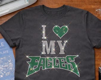 May include: Dark gray t-shirt with the phrase "I ♡ MY EAGLES" in sparkling rhinestones. The heart and the word "EAGLES" are in green. A blue SVG file and a Cricut machine are in the background.