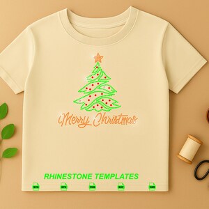 May include: A beige short-sleeved t-shirt featuring a rhinestone graphic of a green Christmas tree with red accents and a star topper. Below the tree, the text Merry Christmas is written in rhinestones. The words RHINESTONE TEMPLATES appear at the bottom.