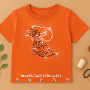 May include: Orange t-shirt with a rhinestone design of a cowboy boot and hat with a rose. The text "RHINESTONE TEMPLATES" is at the bottom of the shirt. A sprig of leaves, scissors, and other craft supplies are arranged around the shirt.