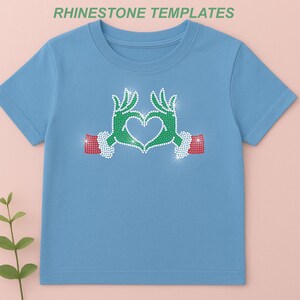 May include: A light blue t-shirt featuring a rhinestone design of green hands forming a heart shape. The hands have red and white accents. The text "RHINESTONE TEMPLATES" is at the top.