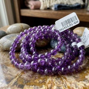 May include: A stretch bracelet composed of round, polished purple amethyst beads. The bracelet features a white rectangular tag with a barcode and the printed text 062453.D1-GM, along with a second white tag partially visible with the text BRUELET.