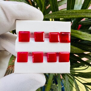 May include: Eight square-cut red gemstones arranged in two rows of four on a white display card. Each gemstone features a faceted surface that reflects light, showcasing a vibrant, uniform red color across all pieces in the collection.