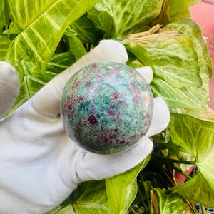 Puede incluir: Una esfera de piedra preciosa pulida, predominantemente verde con inclusiones rojas. La esfera se sostiene en una mano enguantada de blanco, sobre un fondo de exuberante follaje verde. La superficie de la piedra es lisa y reflectante.