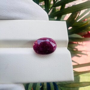 May include: A deep magenta-colored oval gemstone with a faceted surface. The stone displays a translucent quality with internal inclusions and a polished finish, resting on a textured off-white display stand with a horizontal crease.