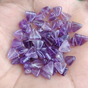 May include: A collection of small, triangular amethyst gemstones. The gemstones are a deep purple colour with translucent areas, and are held in a hand. The stones are likely used for jewellery making or as decorative elements.